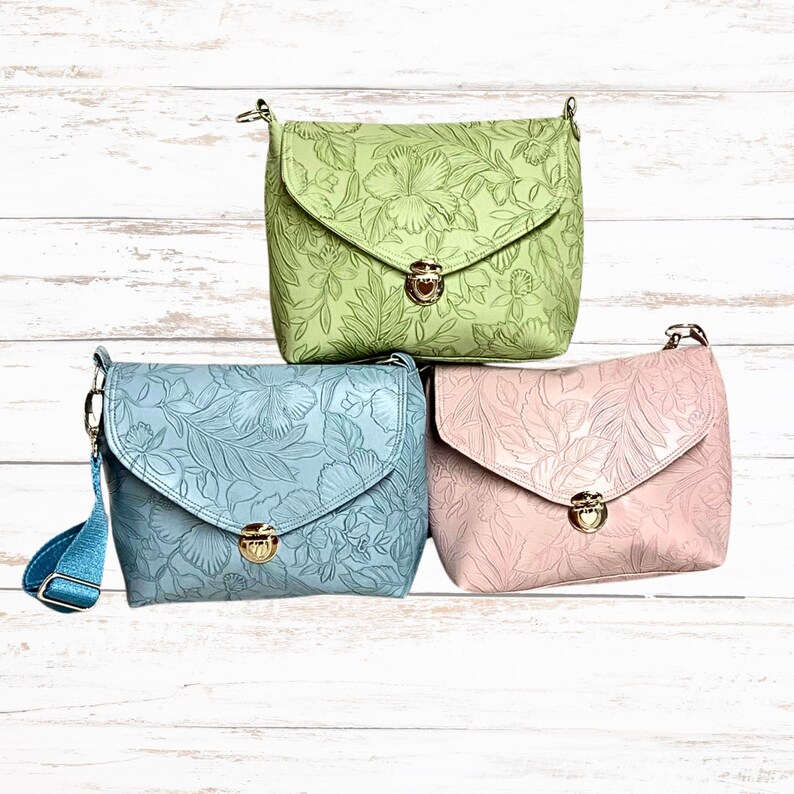May include: Three leather crossbody bags in pastel shades: pale green, light blue, and pink. Each bag has a floral embossed pattern and a flap closure with a gold-tone clasp. The bags have adjustable shoulder straps.