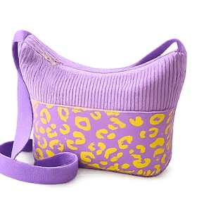 May include: A lavender corduroy crossbody bag with a yellow leopard print design on the lower section. The bag has a long, adjustable strap and a zipper closure. The bag is made of a soft, textured fabric.