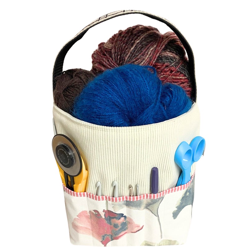 May include: A white fabric tote bag with a floral print and a black handle. The bag is filled with yarn in shades of blue, brown, and red. The bag also contains a pair of scissors, a tape measure, and crochet hooks.