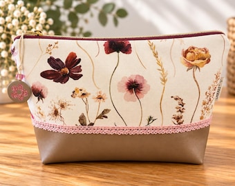 May include: A zippered pouch featuring a cream-colored fabric upper with a floral print of dark red, pink, and yellow wildflowers. The lower section is solid taupe, separated by a pink lace trim. A small circular charm with a pink flower hangs.