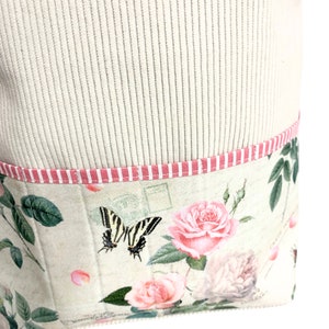 May include: A beige fabric tote bag with a pink and white striped trim. The bottom of the bag features a floral print with pink roses, green leaves, and a black and white butterfly.