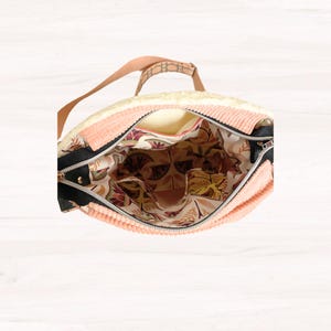 May include: Open view of a peach-colored corduroy tote bag with a black zipper and a patterned interior. The bag has a cream-colored top and a brown strap. The interior features a floral design with shades of brown, green, and peach.