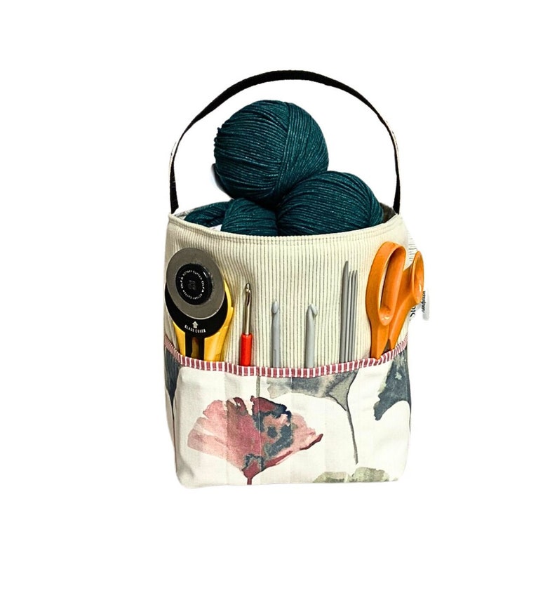 May include: A white canvas tote bag with a floral print and a pink flower design. The bag has a black handle and is filled with balls of yarn, knitting needles, and a pair of scissors.