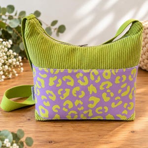 May include: A lime green corduroy hobo bag featuring a wide horizontal band with a purple and lime green leopard print pattern. The bag includes a matching lime green shoulder strap and a silver-toned zipper closure along the top edge.