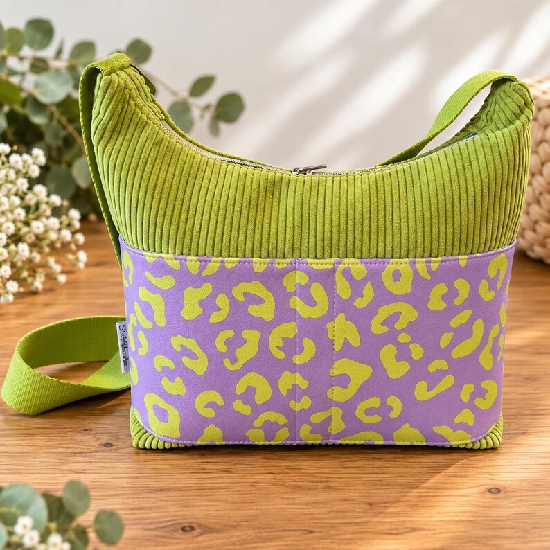 May include: A lime green corduroy hobo bag featuring a wide horizontal band with a purple and lime green leopard print pattern. The bag includes a matching lime green shoulder strap and a silver-toned zipper closure along the top edge.