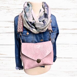 May include: A pale pink crossbody bag with a floral embossed pattern, a gold clasp, and a patterned strap. The bag is worn with a denim jacket and a floral scarf. The strap features a red and white geometric design.