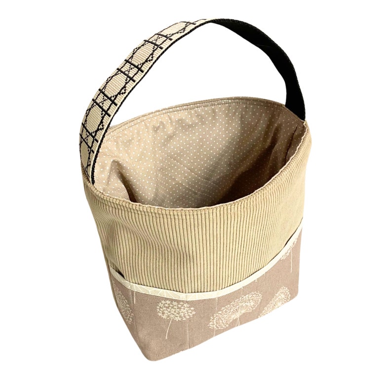 May include: A beige fabric storage basket with a black and white patterned handle. The basket has a beige and white floral print on the bottom and a beige corduroy fabric on the top.
