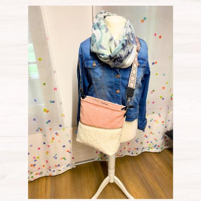 May include: A denim jacket, floral scarf, and a pink and white crossbody bag are displayed on a mannequin. The bag has a textured pink top and a fluffy white bottom. The bag strap has a geometric pattern.
