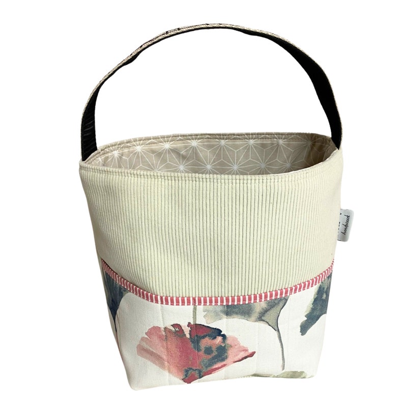 May include: A beige fabric storage basket with a black handle and a floral print on the bottom. The basket has a red and white stripe around the top edge. The basket is labelled "handmade".