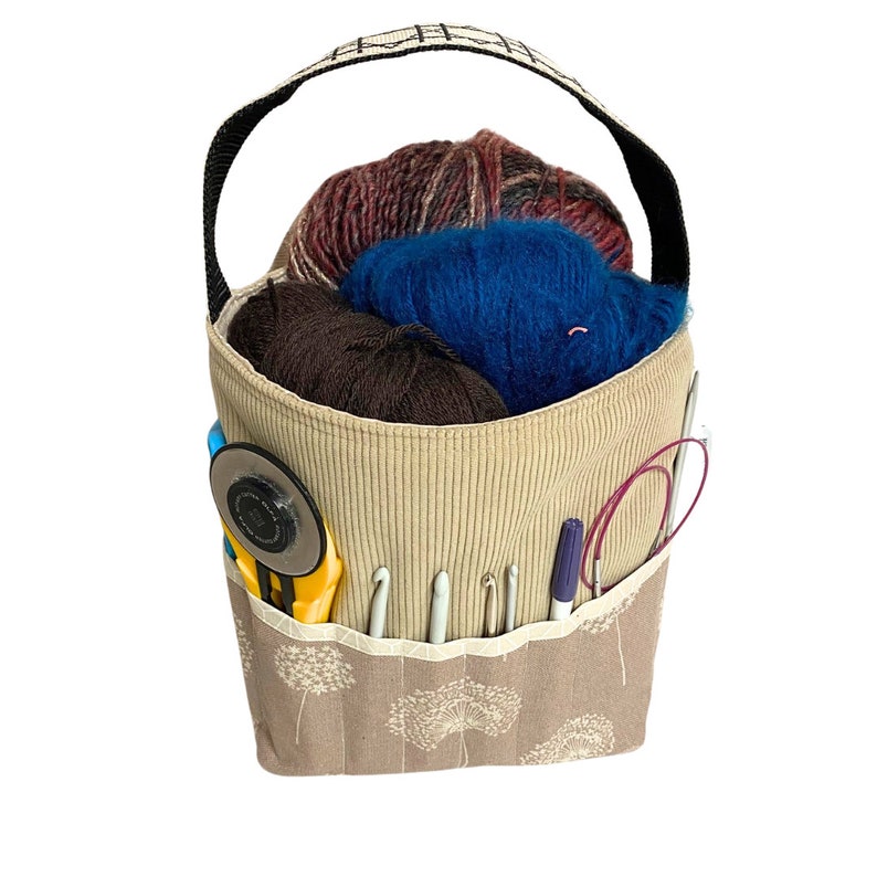 Puede incluir: Una bolsa de tela beige con un patr&oacute;n de diente de le&oacute;n marr&oacute;n y blanco. La bolsa tiene un asa negra y est&aacute; llena de hilo, agujas de tejer y otros materiales de artesan&iacute;a.