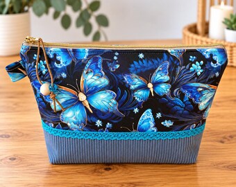 May include: A zippered pouch featuring a black fabric top with a vibrant blue butterfly and floral pattern. The bottom section is made of solid grey ribbed fabric, separated by a teal lace trim. A gold zipper pull includes a leather cord and bead.