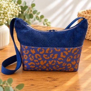 May include: A crossbody bag featuring a dark blue corduroy upper section and a lower section with a blue and orange leopard print pattern. The bag includes a matching blue shoulder strap and a zippered closure along the top edge of the corduroy.