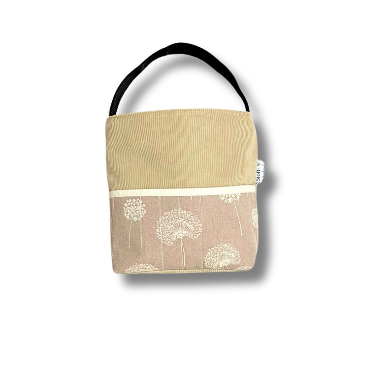 May include: A small beige tote bag with a black handle. The bag has a beige corduroy top and a bottom with a white dandelion pattern on a light pink background. The bag has a white label with the text "Stoff & Stil" on the side.