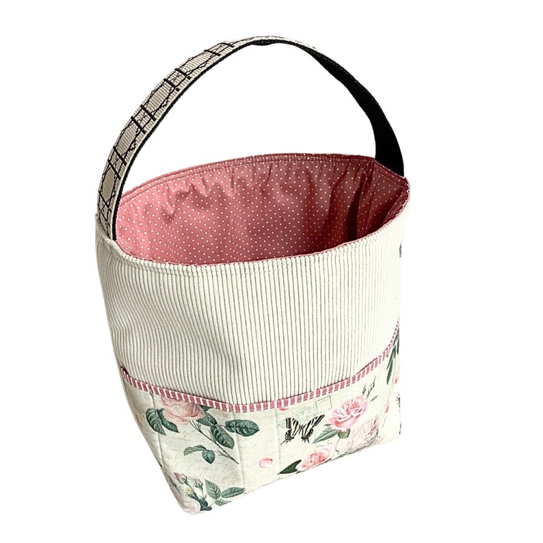May include: A white and pink fabric storage basket with a black handle and floral print. The basket has a pink polka dot lining and multiple pockets.
