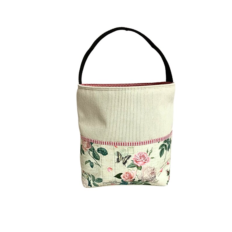 May include: A white and green fabric tote bag with a black handle. The bag has a floral print with pink roses and butterflies. The bag has a red and white striped trim.