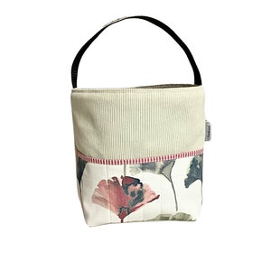 May include: A beige and white fabric tote bag with a black handle. The bag features a floral print with pink, green, and grey flowers. The bag has a red and white striped trim. The bag has a small white label with the word "handmade" on it.