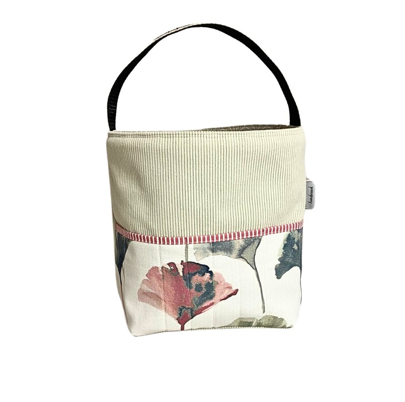 May include: A beige and white fabric tote bag with a black handle. The bag features a floral print with pink, green, and grey flowers. The bag has a red and white striped trim. The bag has a small white label with the word "handmade" on it.