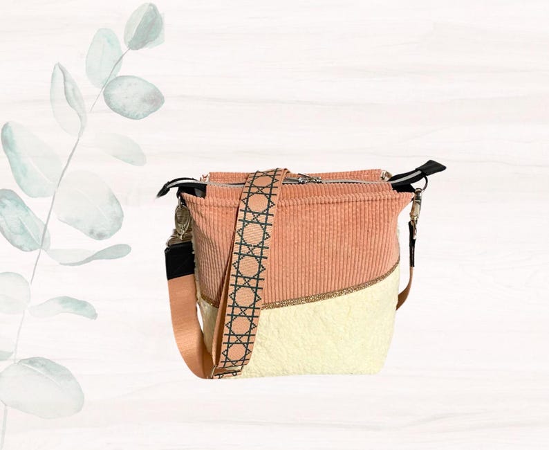 May include: A peach and cream-colored crossbody bag with a patterned strap. The bag features a corduroy texture on the top half and a smooth, cream-colored bottom half. The strap has a geometric design in black and tan.