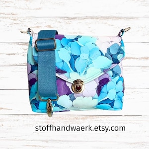May include: A floral print crossbody bag with a teal adjustable strap and gold-tone hardware. The bag features a flap closure with a twist lock. The floral pattern includes shades of blue, purple, and white. The Etsy shop name is visible on the bottom.