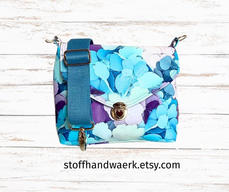 May include: A floral print crossbody bag with a teal adjustable strap and gold-tone hardware. The bag features a flap closure with a twist lock. The floral pattern includes shades of blue, purple, and white. The Etsy shop name is visible on the bottom.