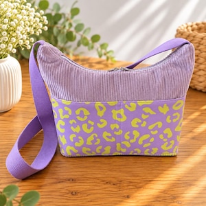 May include: A lavender corduroy shoulder bag featuring a horizontal zipper closure and a wide purple fabric strap. The lower section of the bag is made of a smooth purple fabric with a bright lime green leopard print pattern across the front.