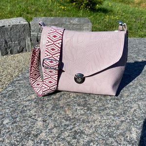May include: A pale pink crossbody bag with a floral embossed pattern. The bag has a patterned strap in white and burgundy. It features a silver clasp closure and is on a stone surface.
