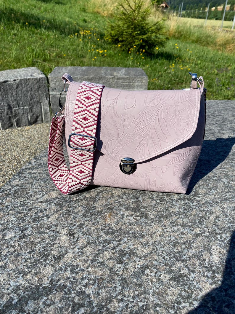 May include: A pale pink crossbody bag with a floral embossed pattern. The bag has a patterned strap in white and burgundy. It features a silver clasp closure and is on a stone surface.