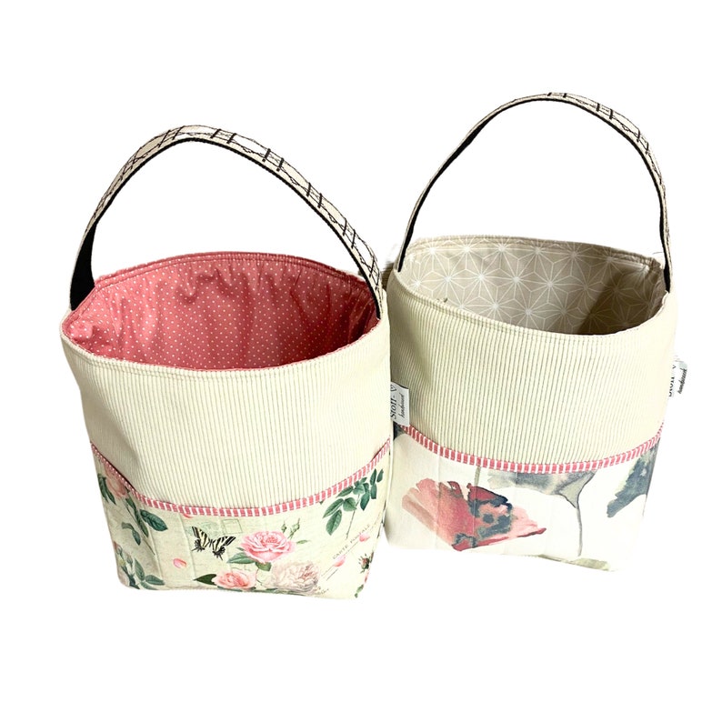May include: Two cream-coloured fabric storage baskets with black handles. The baskets have a floral print and a pink and white polka dot lining.