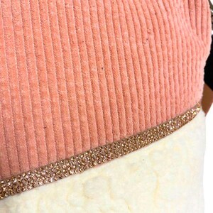 May include: Close-up of a Christmas stocking with a peach-colored corduroy top and a white, fluffy bottom. A band of gold-colored rhinestones separates the two sections. The stocking is likely for holiday decor.