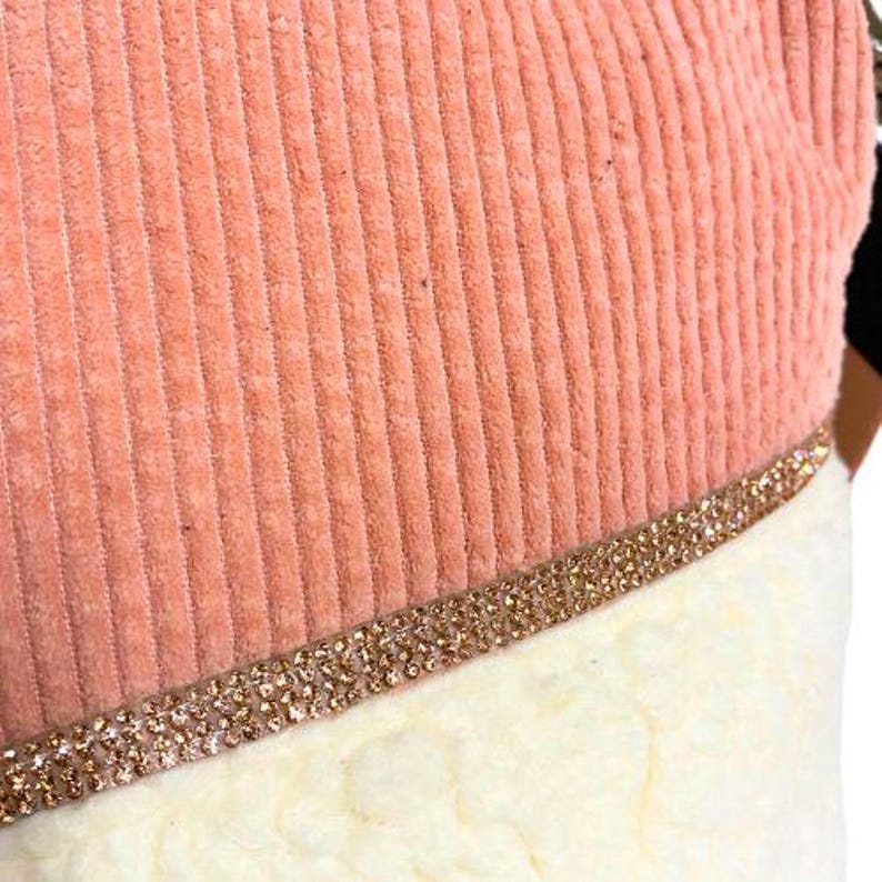 May include: Close-up of a Christmas stocking with a peach-colored corduroy top and a white, fluffy bottom. A band of gold-colored rhinestones separates the two sections. The stocking is likely for holiday decor.
