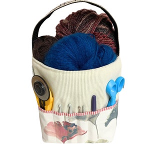 May include: A white fabric tote bag with a floral print and a handle. The bag is filled with yarn in shades of blue, brown, and red. There are also knitting needles and scissors in the bag.