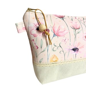 May include: A pink zippered pouch with a floral print and a beige tassel. The pouch has a white crocheted trim and a beige fabric lining.