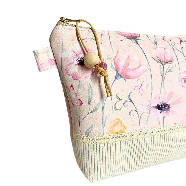 May include: A pink zippered pouch with a floral print and a beige tassel. The pouch has a white crocheted trim and a beige fabric lining.
