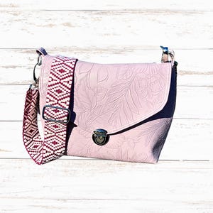 May include: A pale pink crossbody bag with a floral embossed pattern. It has a patterned strap in white and burgundy. The bag features a flap closure with a silver-tone clasp and hardware.