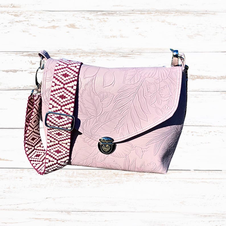 May include: A pale pink crossbody bag with a floral embossed pattern. It has a patterned strap in white and burgundy. The bag features a flap closure with a silver-tone clasp and hardware.