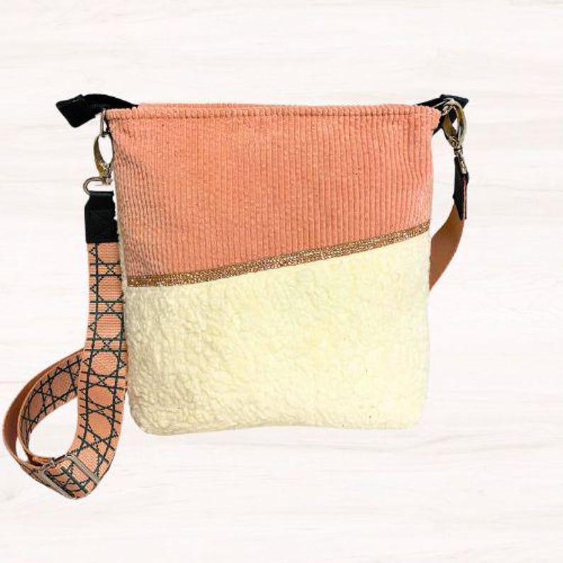 May include: A two-tone crossbody bag with a peach corduroy top and a cream-colored, textured bottom. A gold-colored band with small embellishments separates the two sections. The bag has a patterned strap and black accents.