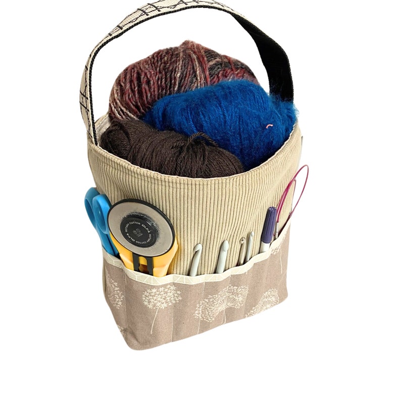 May include: A beige canvas tote bag with a floral pattern and multiple pockets. The bag is filled with yarn, knitting needles, and a pair of scissors.
