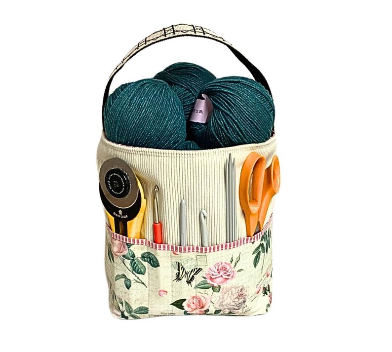 May include: A white canvas tote bag with a black handle and floral print. The bag is filled with green yarn balls, scissors, and knitting needles.