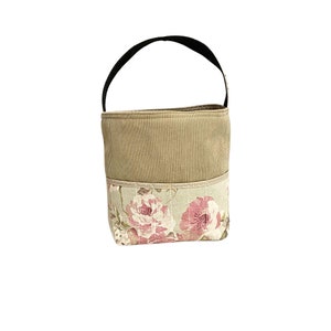 Puede incluir: Una peque&ntilde;a bolsa de tela beige con un asa negra. La bolsa tiene un estampado floral con rosas rosas en la mitad inferior.