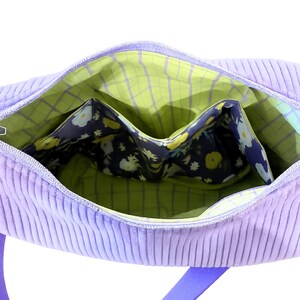 May include: Open view of a lavender corduroy tote bag with a blue strap. The interior features a green plaid lining and a floral print pocket. The bag is designed with multiple compartments for organization.