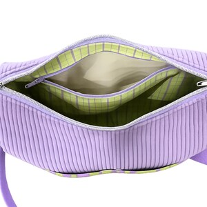 May include: Open view of a lavender corduroy handbag with a silver zipper. The interior features a light beige lining and a green and white plaid pocket. The bag has a matching lavender handle.