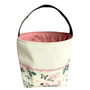 May include: A white fabric tote bag with a pink polka dot lining and a black handle. The bag has a floral print with roses and butterflies.