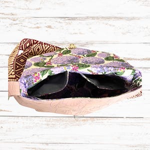 May include: A floral shoulder bag with a light pink embossed leather exterior and a patterned strap. The bag's interior features a floral print with purple hydrangeas and black pockets. The strap has a geometric design in burgundy and tan.