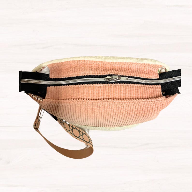 May include: A peach-colored corduroy fanny pack with a black zipper and a tan strap. The bag has a cream-colored lining and a geometric pattern on the strap. The zipper is silver.
