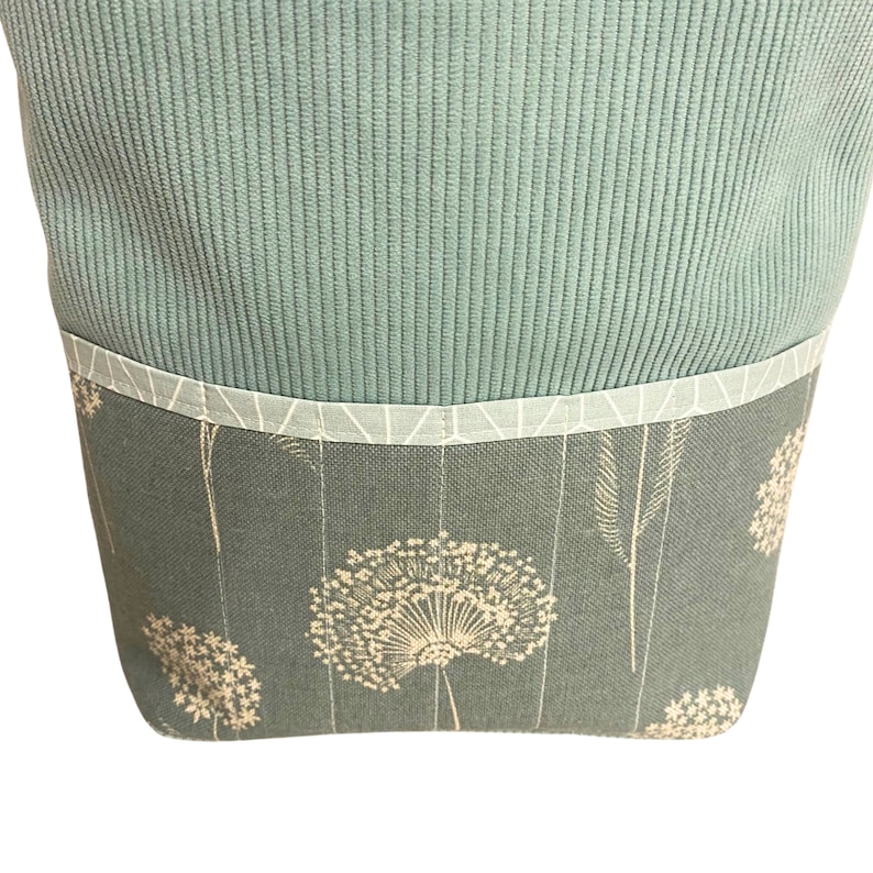 Puede incluir: Una bolsa de tela azul verdosa con un estampado floral blanco. La bolsa tiene una solapa superior con un ribete blanco.