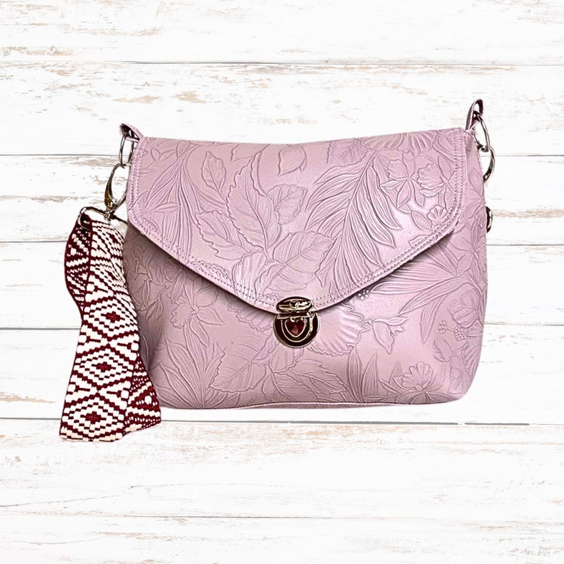 May include: A lilac crossbody bag with a floral embossed pattern. It has a flap closure with a silver clasp and chain strap. A patterned strap in burgundy and white is attached to the side.