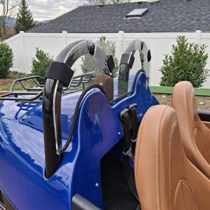 May include: A close-up of a blue roadster's interior. The vehicle features tan leather seats with white stitching, black roll bars, and a clear windshield. The exterior is a vibrant blue, with black accents.