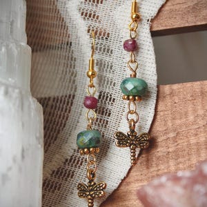 May include: A pair of gold-toned drop earrings featuring a dragonfly charm at the base, a faceted teal bead, and a small round red bead. The earrings are suspended from gold-toned fishhook wires, showcasing a delicate metal construction.