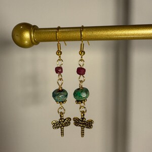 May include: A pair of dangle earrings with gold-colored hooks and chains. Each earring features a dragonfly charm, a faceted teal bead, and small red beads. The earrings are displayed on a gold-colored rod.