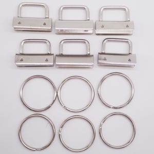 May include: A collection of silver-toned metal hardware components, including rectangular clasps with arched handles and circular key rings. The clasps have small triangular holes. The items are arranged on a white surface.